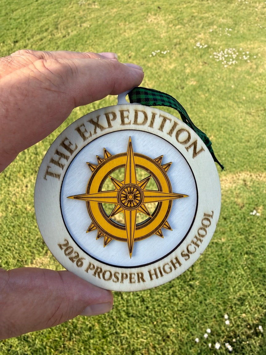 Laser-engraved wood ornament reading 'THE EXPEDITION' and '2026 PROSPER HIGH SCHOOL', held in hand over grass to show its size