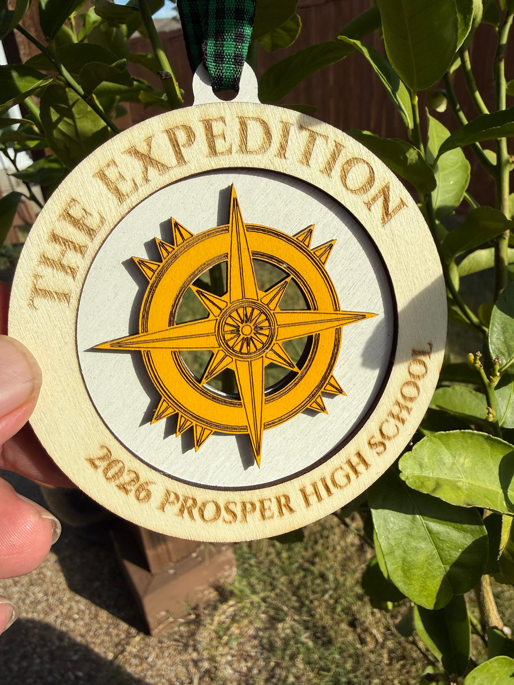Laser-engraved wood ornament reading 'THE EXPEDITION' and '2026 PROSPER HIGH SCHOOL', held up in front of lemon-tree leaves